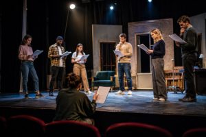 Troupe de théâtre amateur en pleine répétition sur scène sous les projecteurs, guidée par un metteur en scène dans une salle de spectacle